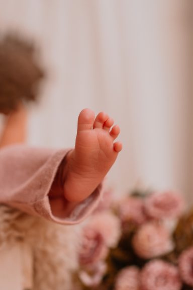 Babyfuß in einem weichen, rosa Outfit, im Vordergrund sind Blumen zu sehen.