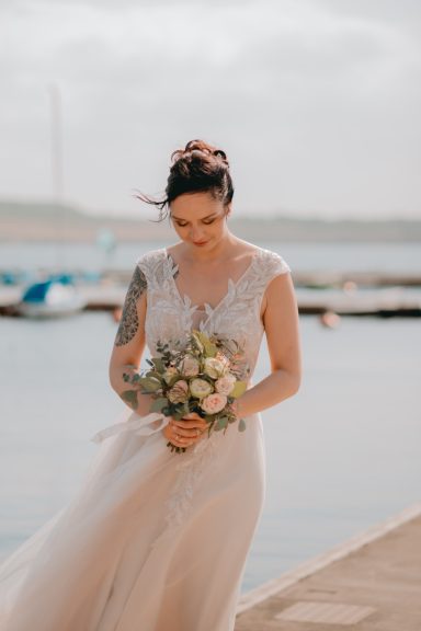 Braut mit Blumenstrauß in einem eleganten Kleid am Wasser, sanfte Brise weht.