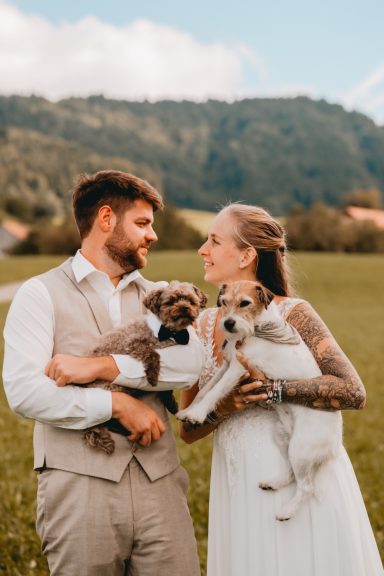 Braut und Bräutigam halten zwei Hunde vor einer ländlichen Kulisse.