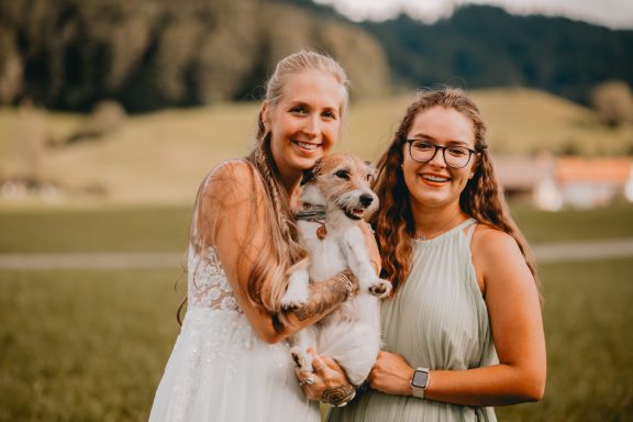 Zwei Frauen im Freien, eine mit einem Hund, auf einer grünen Wiese vor Bergen.