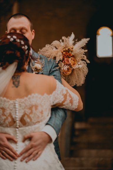 Braut und Bräutigam umarmen sich, Braut trägt einen Blumenstrauß und ein elegantes Kleid.
