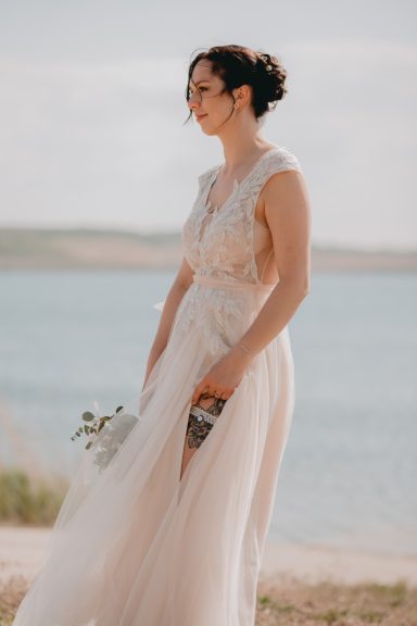 Braut in einem eleganten, fließenden Kleid am Ufer eines Gewässers, mit Blumenstrauß.