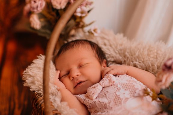 Schlafendes Baby in zarter rosa Kleidung, umgeben von Blumen und weichem Stoff.