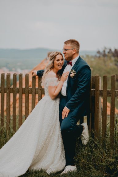 Hochzeitspaar posiert lächelnd vor einem Holzzaun mit Landschaft im Hintergrund.