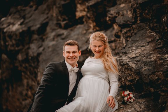 Hochzeitsfoto eines Paares auf einem Felsen, lächelnd mit Blumenstrauß.