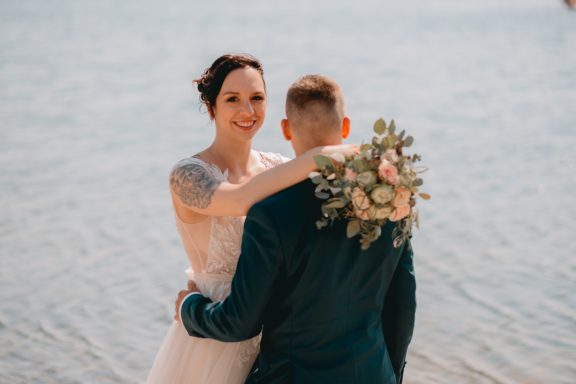 Braut und Bräutigam stehen am Wasser, umarmt, mit einem Blumenstrauß im Hintergrund.