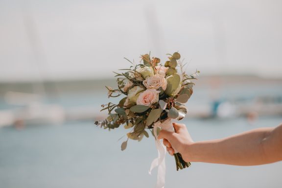 Hochzeitsstrauß mit rosa Blumen und grünem Blattwerk vor einem maritimen Hintergrund.