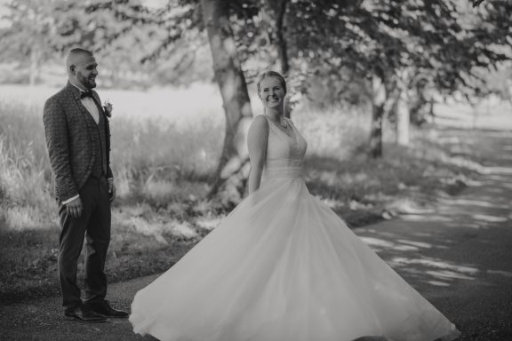 Braut in einem weißen Kleid, steht auf einem Weg, Brautigam im Hintergrund.