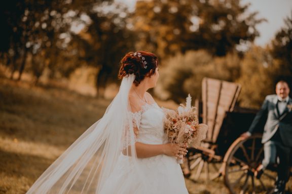 Braut mit Brautstrauß im Freien, im Hintergrund eine Hochzeitskutsche und ein Bräutigam.