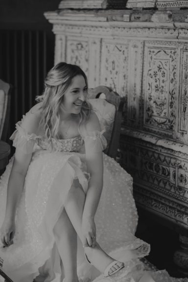 Lächelnde Frau in einem eleganten Hochzeitskleid sitzt und zieht ihren Schuh an.