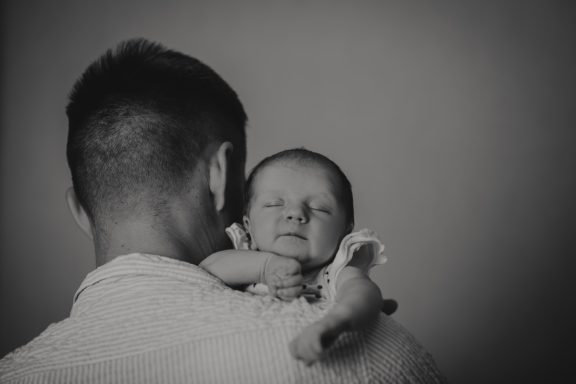 Vater hält lächelnd ein Baby im Arm, beide von hinten zu sehen.