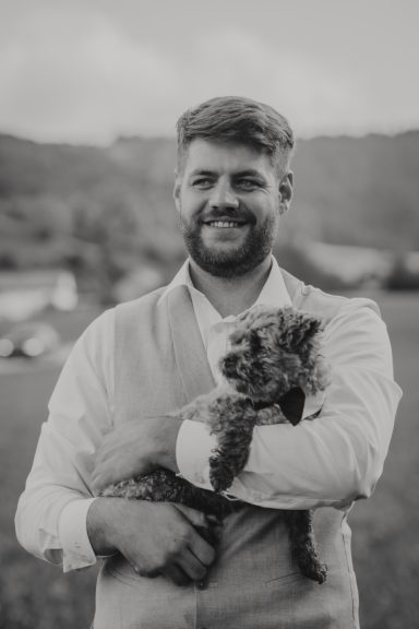 Ein lächelnder Mann in Weste hält einen kleinen Hund auf dem Arm, mit Natur im Hintergrund.