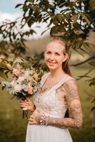 Braut mit Blumenstrauß, tätowierten Armen, lächelnd vor natürlicher Kulisse.