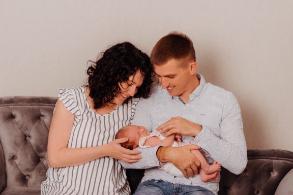 Junge Eltern halten ihr neugeborenes Baby, lächelnd auf einem Sofa.