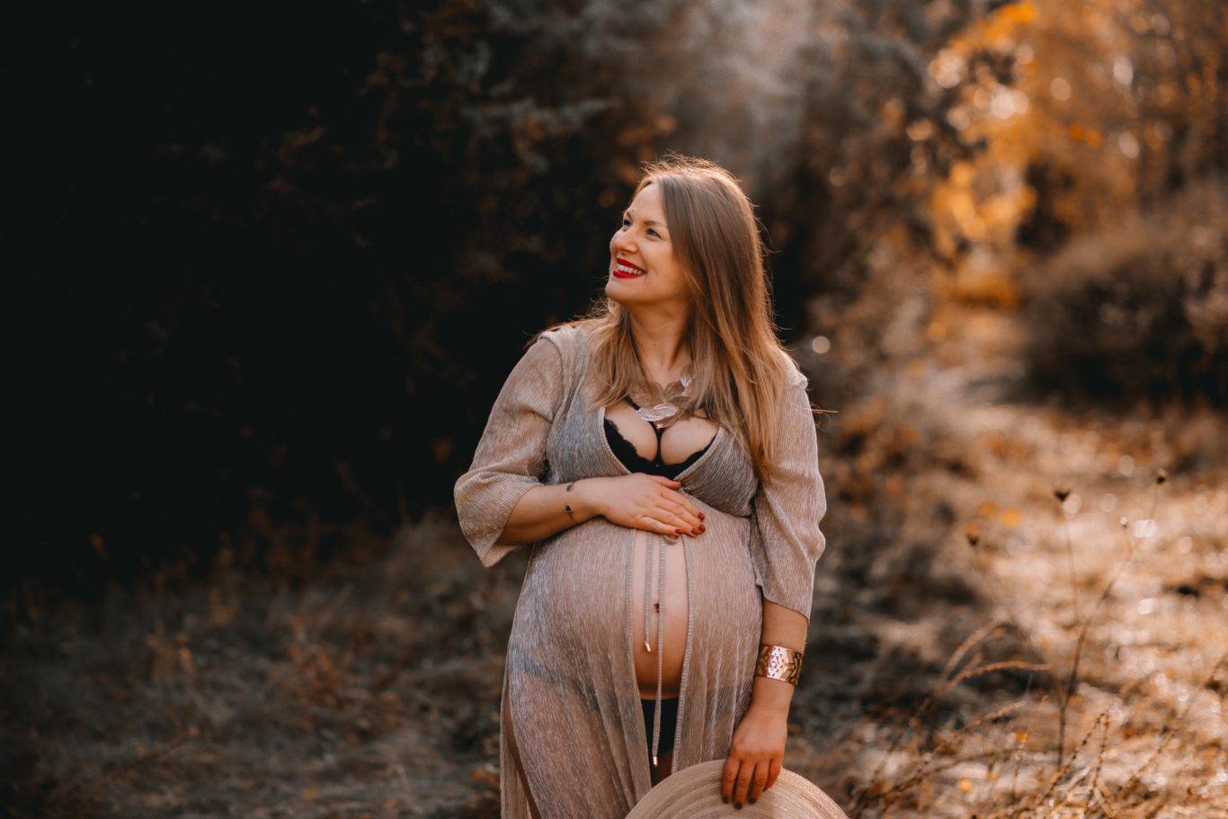 Schwangere Frau mit Lächeln, trägt ein leichtes Kleid in einer natürlichen Umgebung.