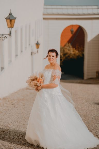 Braut in einem weißen Kleid mit Blumenstrauß, steht vor einer sonnigen Kulisse.