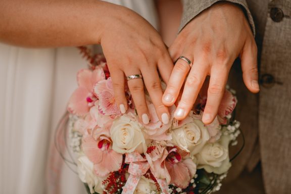 Hände mit Ringen halten einen Blumenstrauß in Pastelltönen.