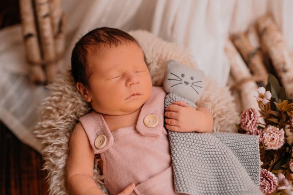 Neugeborenes Baby schläft mit einer Spielzeugkatze in der Hand, umgeben von Blumen.