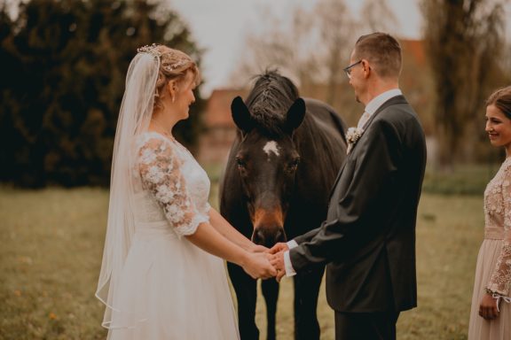 Braut und Bräutigam halten Händchen, während ein schwarzes Pferd ihnen gegenüber steht.